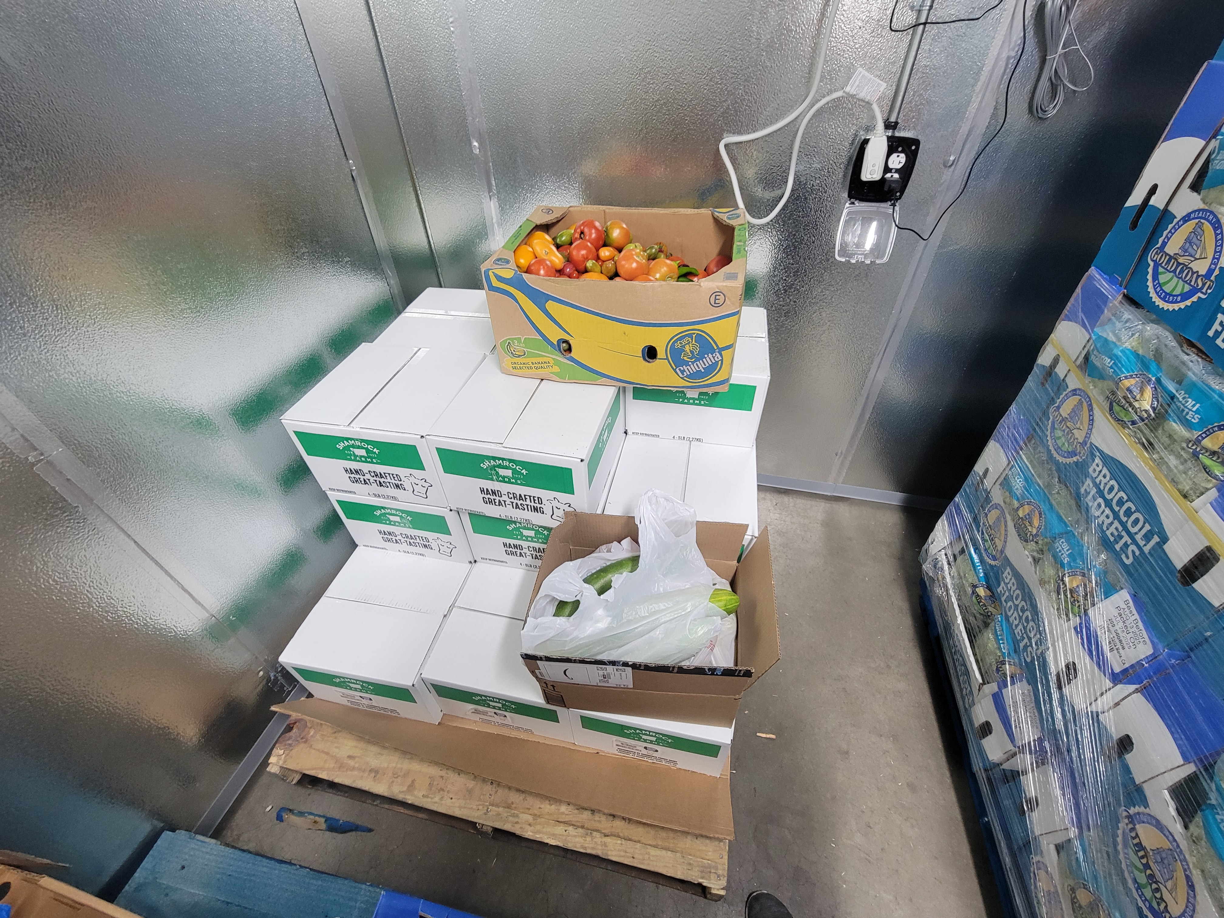 Banana box of colorful tomatoes and cardboard box of zucchinis on top of boxes of milk on a pallet in a fridge box unit.